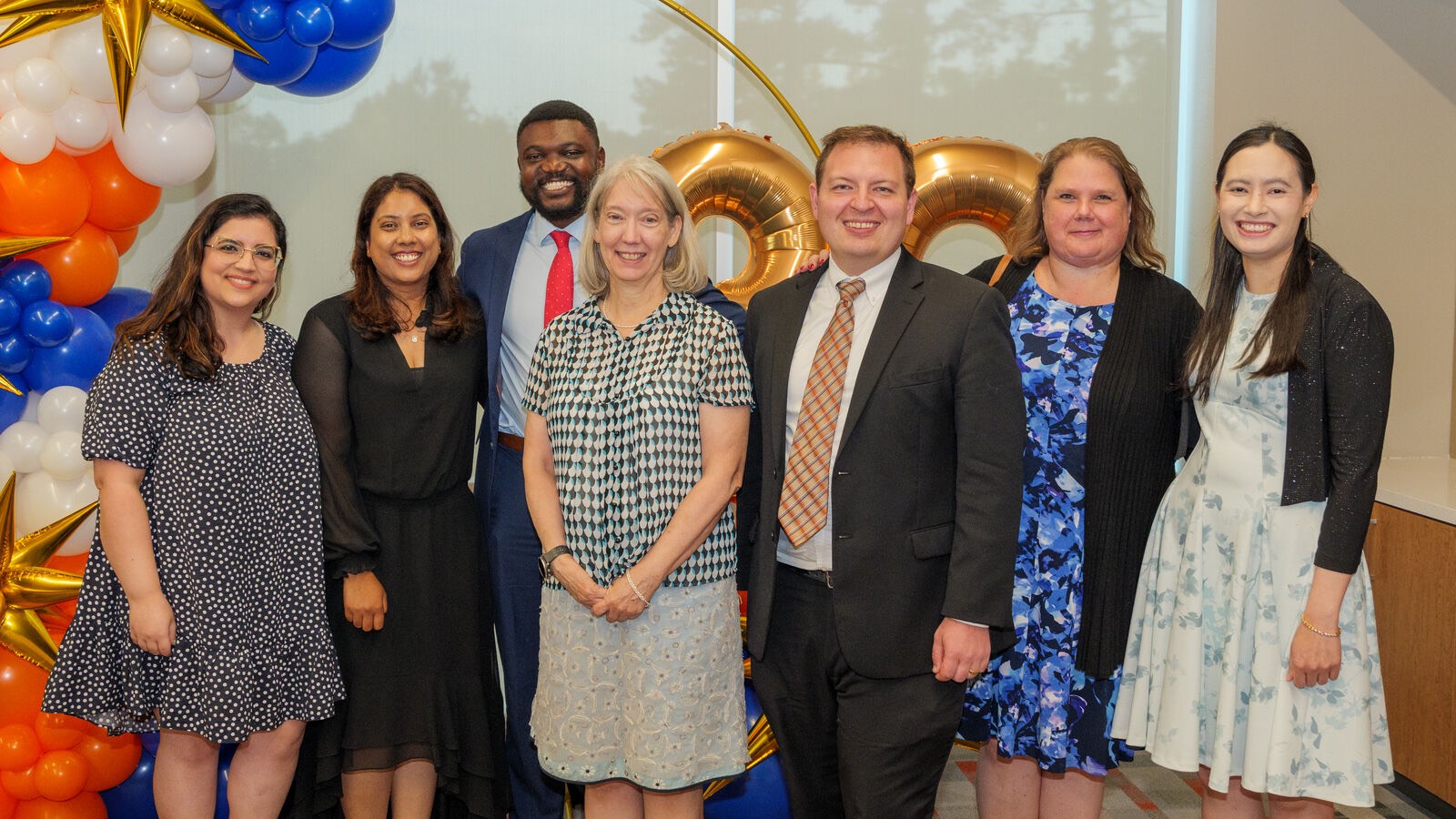 "Group photo of seven individuals dressed formally, standing in front of a colorful balloon arch featuring blue, orange, white, and gold balloons. Gold number balloons are visible in the background, and a window with trees outside adds natural light to the setting.