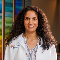 Woman with long, curly brown hair and brown eyes smiling. Woman is wearing a doctor's white coat. 