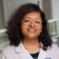 A woman with curly dark hair wearing rectangular glasses and a white lab coat smiles warmly. She is standing in an indoor environment with a softly blurred background