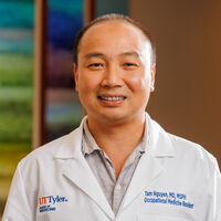 A smiling man wearing a white lab coat with 'UT Tyler' and his name embroidered on it, standing indoors with a colorful, blurred background