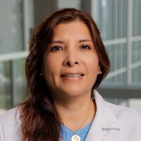 A woman with long, dark brown hair smiles gently. She is wearing a white lab coat over a blue shirt, with a neutral gray background behind her.