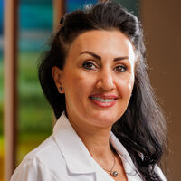 A woman with long, dark hair smiles warmly. She is wearing a white lab coat, with a colorful background behind her