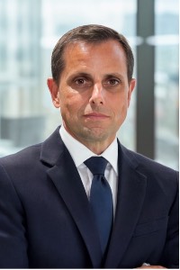 man with dark hair, tan, in a dark navy suit with navy tie, posing for a photo.